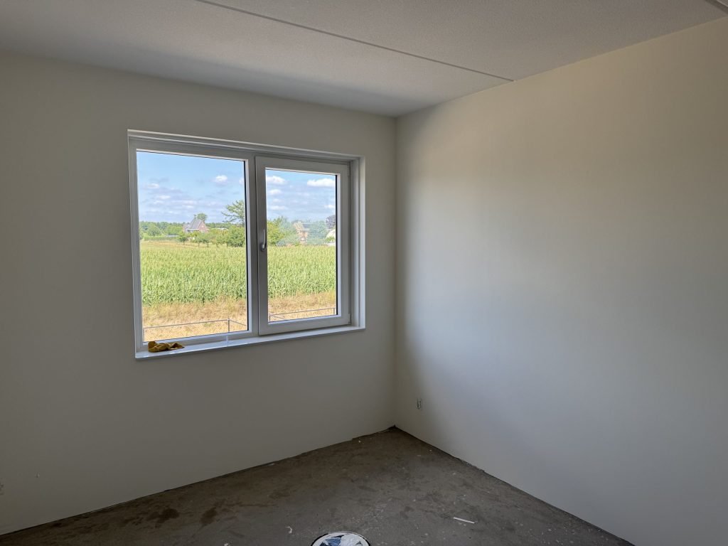 Slaapkamer in een nieuwe woning met kale witte wanden en uitzicht op een maisveld